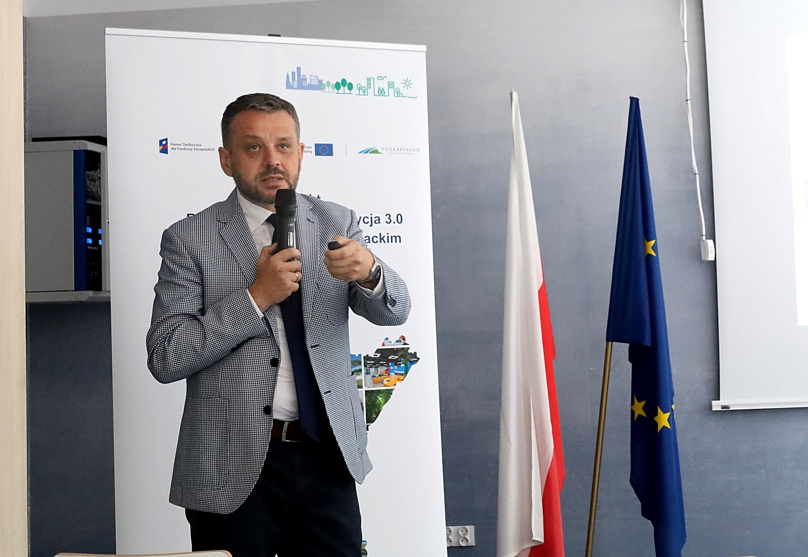 Mężczyzna (Andrzej Brzozowy) w szarym garniturze trzyma mikrofon i wskaźnik podczas przemówienia na konferencji. Za nim znajduje się flaga Polski, flaga UE i baner z tekstem i grafiką.
